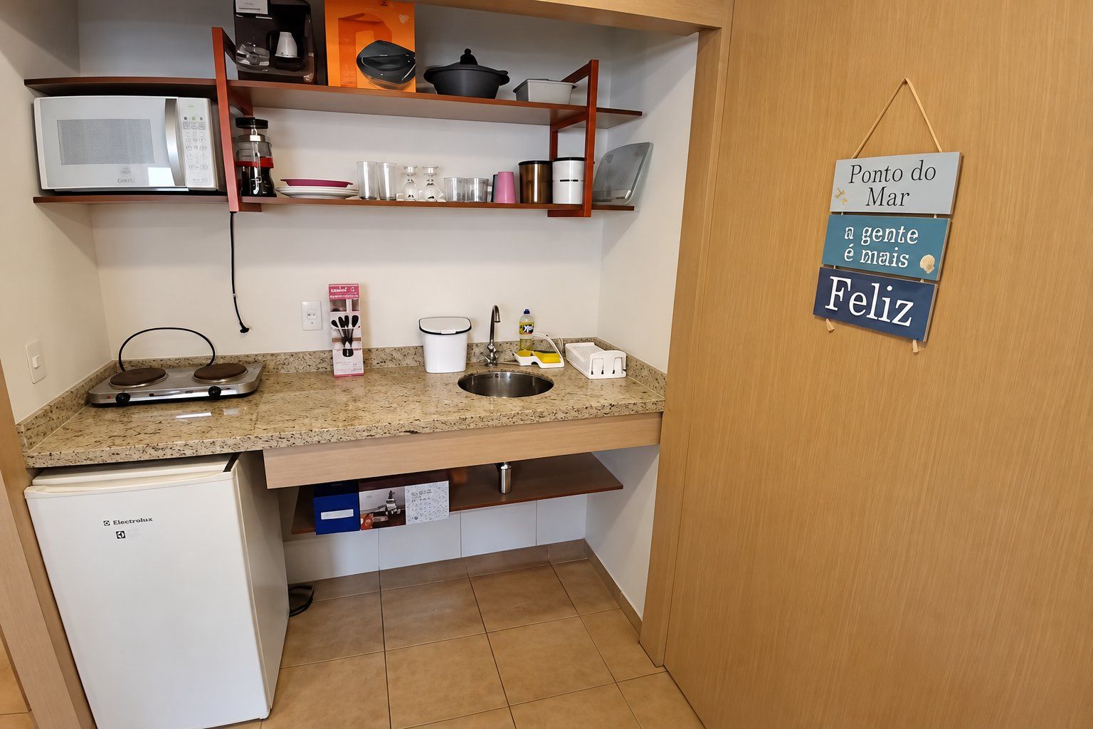 Une cuisine compacte équipée d’un four à micro-ondes, d’un évier et d’un petit réfrigérateur. Une étagère au-dessus du plan de travail permet de ranger divers ustensiles de cuisine. Un panneau avec un message est accroché au mur.