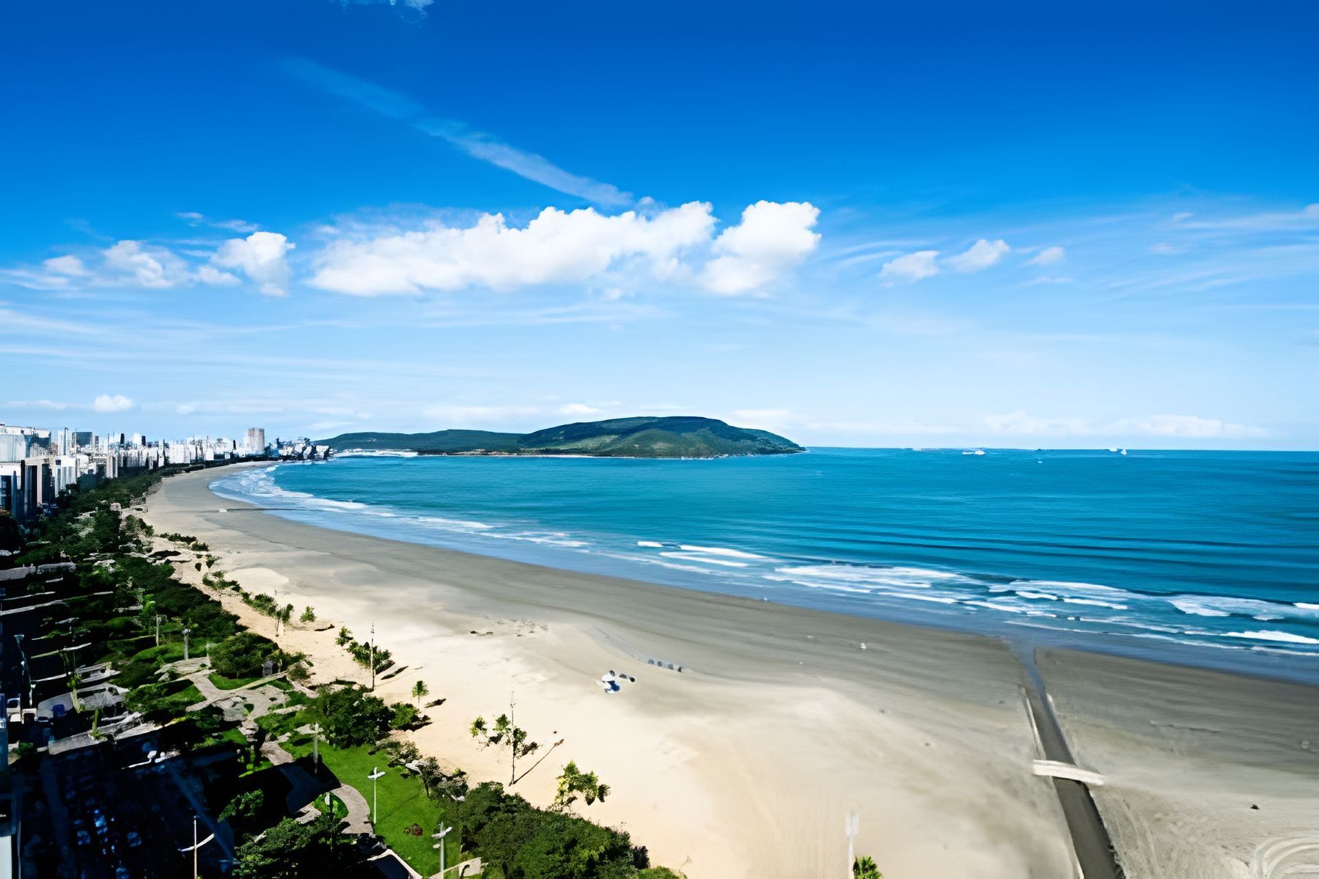 Paisagem costeira urbana com uma praia extensa, uma pequena ilha e um céu azul límpido.