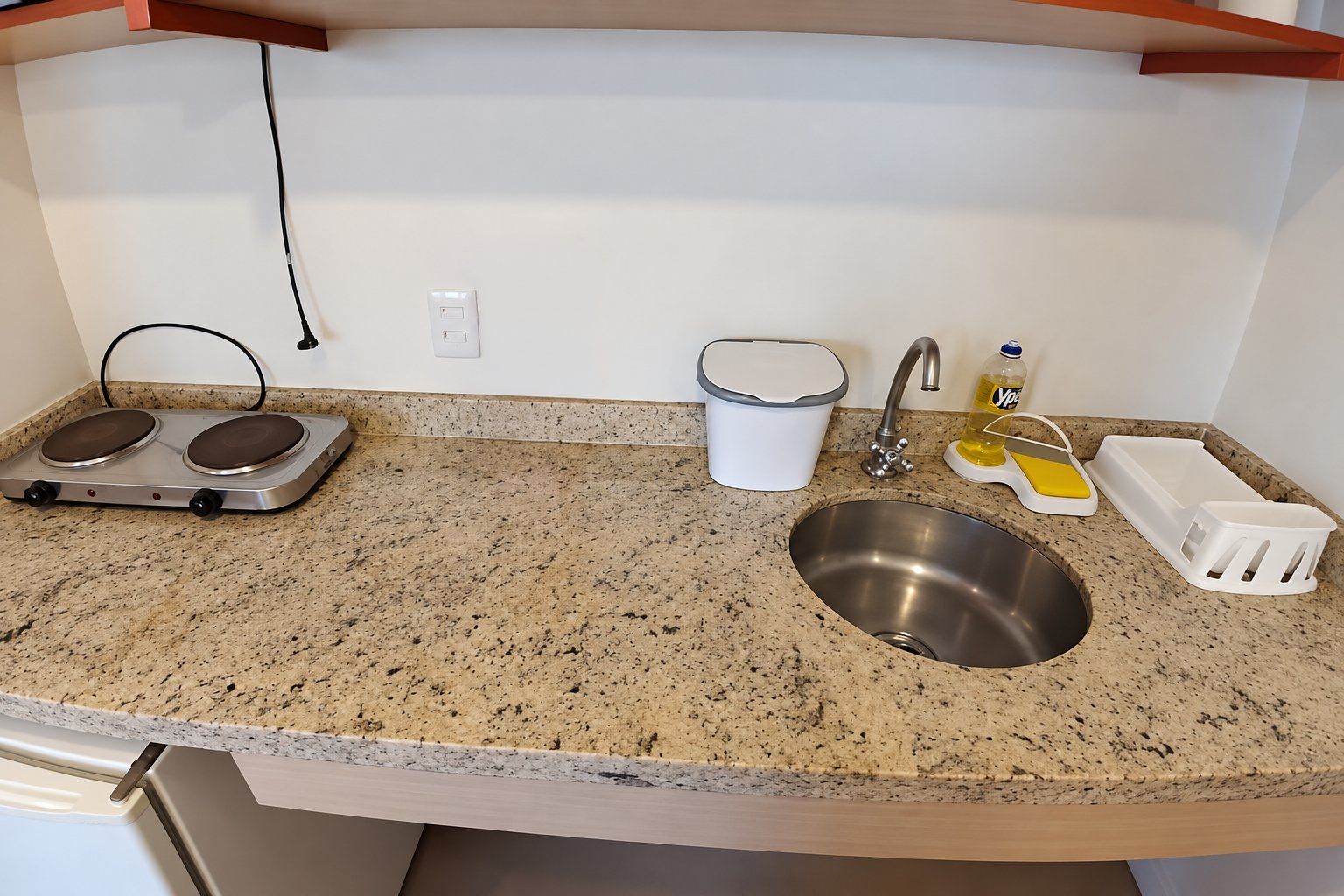 A kitchen with a granite countertop, stainless steel sink, two electric kettles, a white trash bin, a faucet, and a soap dispenser.