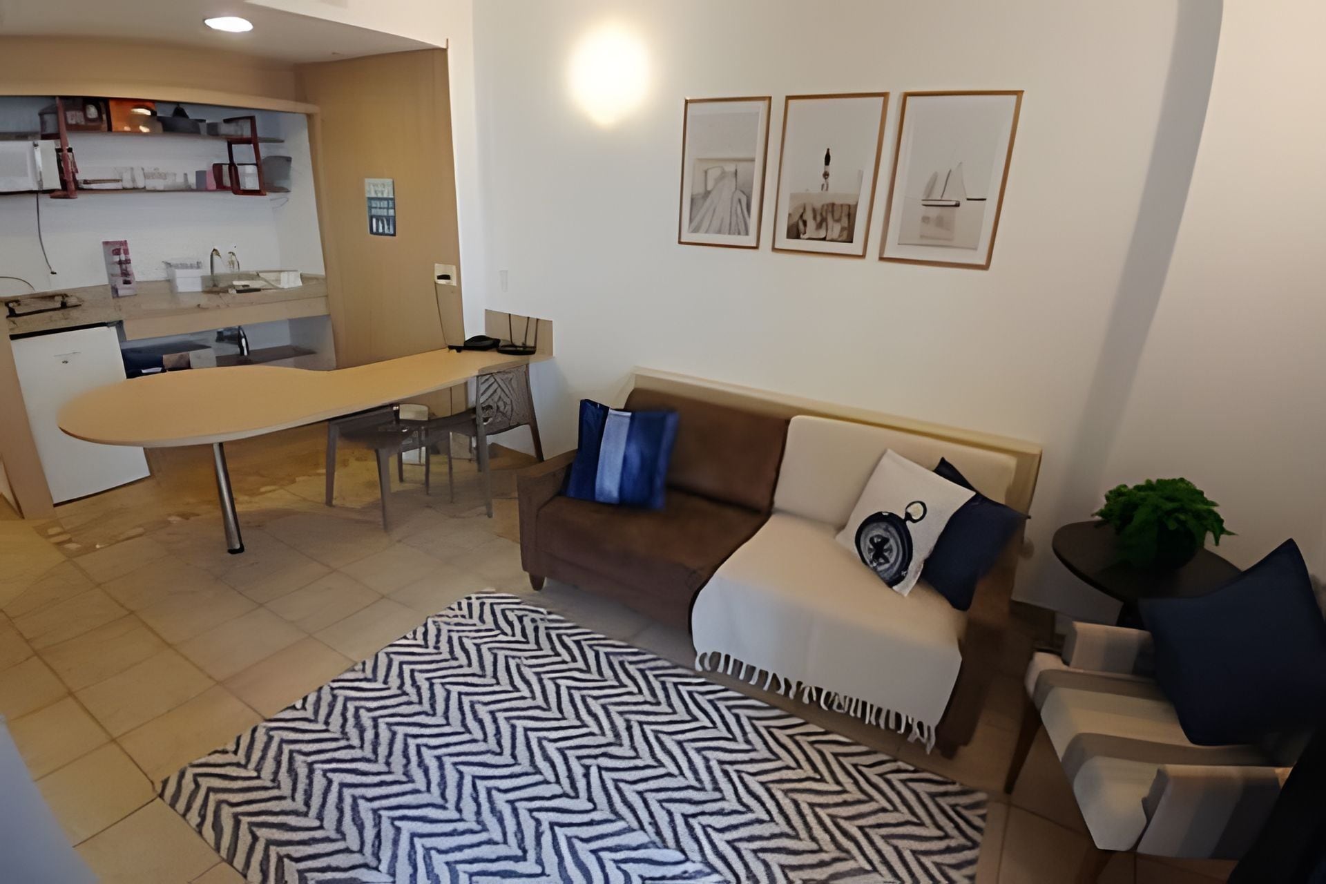 A living room with a sofa, a coffee table, and a patterned rug. There is a kitchen area with a counter and a refrigerator.