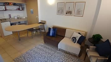 A living room with a sofa, a coffee table, and a patterned rug. There is a kitchen area with a counter and a refrigerator.