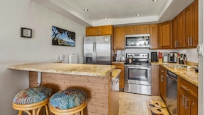 A kitchen with wooden cabinets, stainless steel appliances, a granite countertop, and two stools. - Oceanfront Condo Near Napili Bay 1br - Unit A17 1 Bedroom Condo (Lahaina)