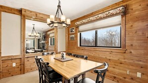 A wooden-paneled dining room with a large table and chairs, a chandelier, and a window with blinds. - Seven Springs Meadowridge 3brloft Premier-cssv8011 3 Bedroom Condo (Champion)