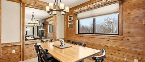 A wooden-paneled dining room with a large table and chairs, a chandelier, and a window with blinds.