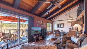 A spacious living area with a large TV, wooden ceiling beams, a dining area, and a view through large sliding doors. - Tahoe City - Lake Forest Condo 3 Bedroom Condo (Tahoe City)