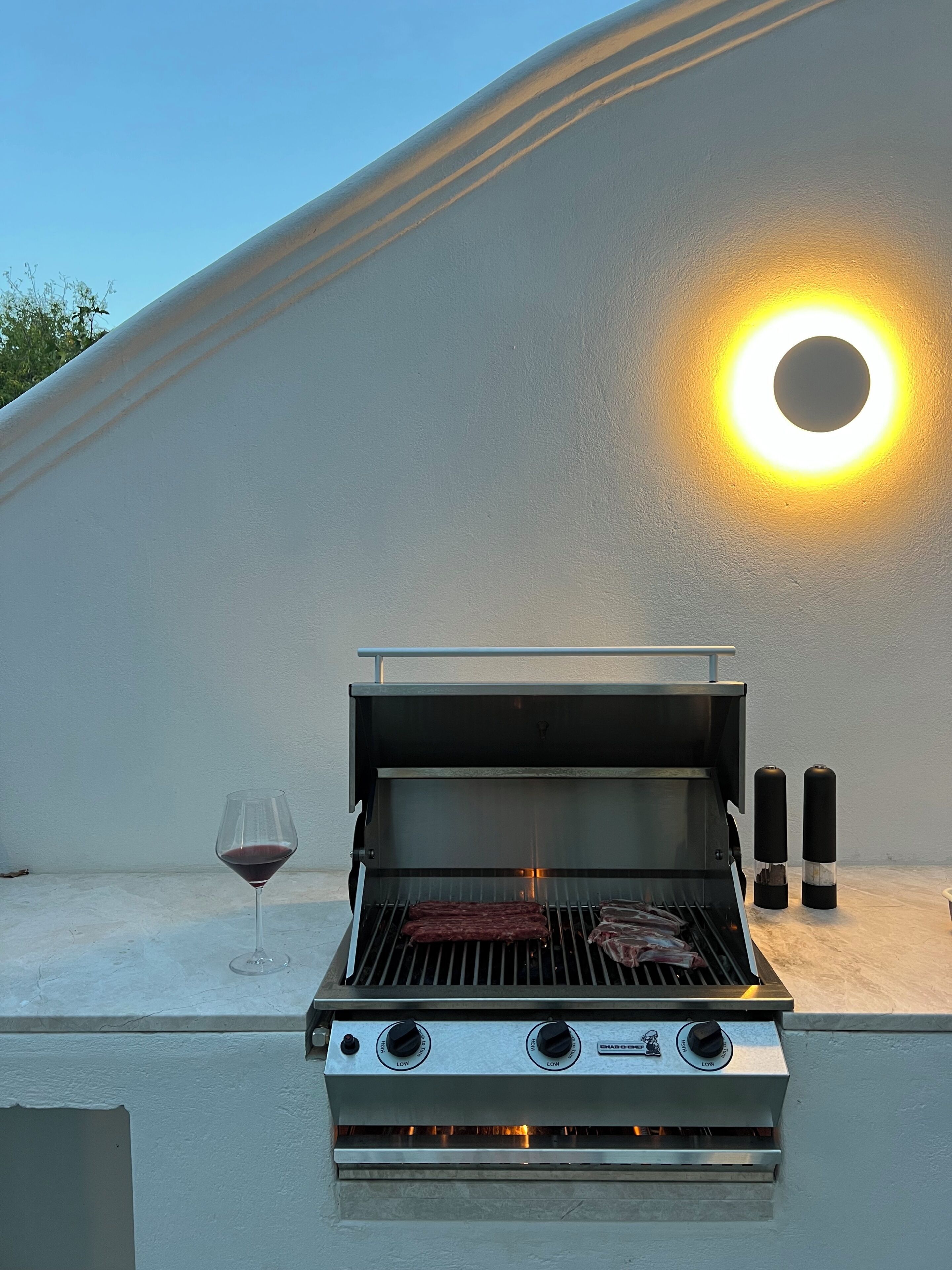 Un barbecue d’extérieur, avec un verre à vin posé à côté, le tout installé devant un mur blanc sur lequel est fixée une lampe circulaire.