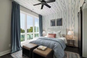 Une chambre avec une grande fenêtre, un ventilateur de plafond, un lit avec une tête de lit, deux tables de chevet avec des lampes et un banc avec un livre.