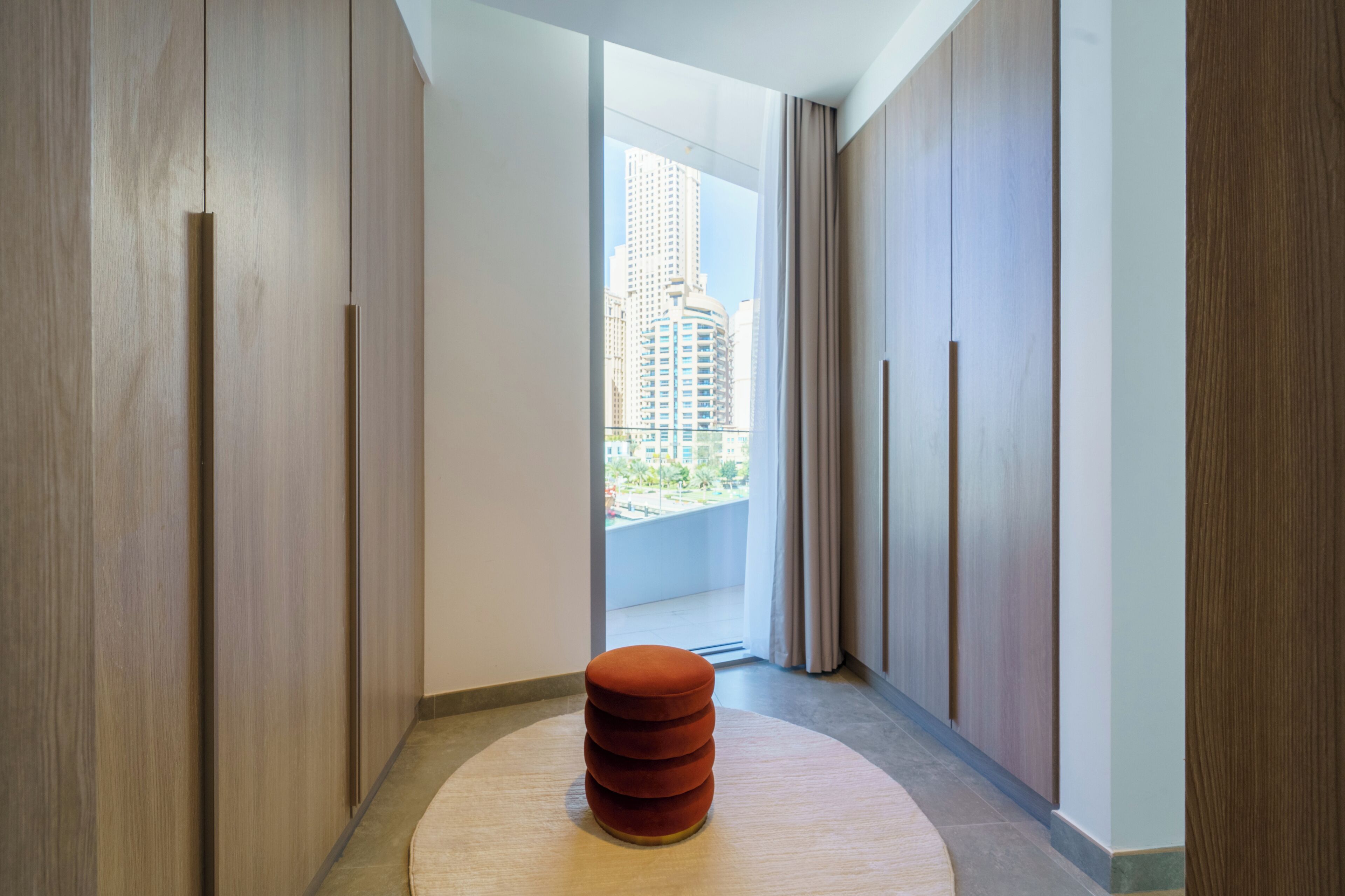 A hallway with a circular rug, wooden wardrobe, and a view of a cityscape through a window.