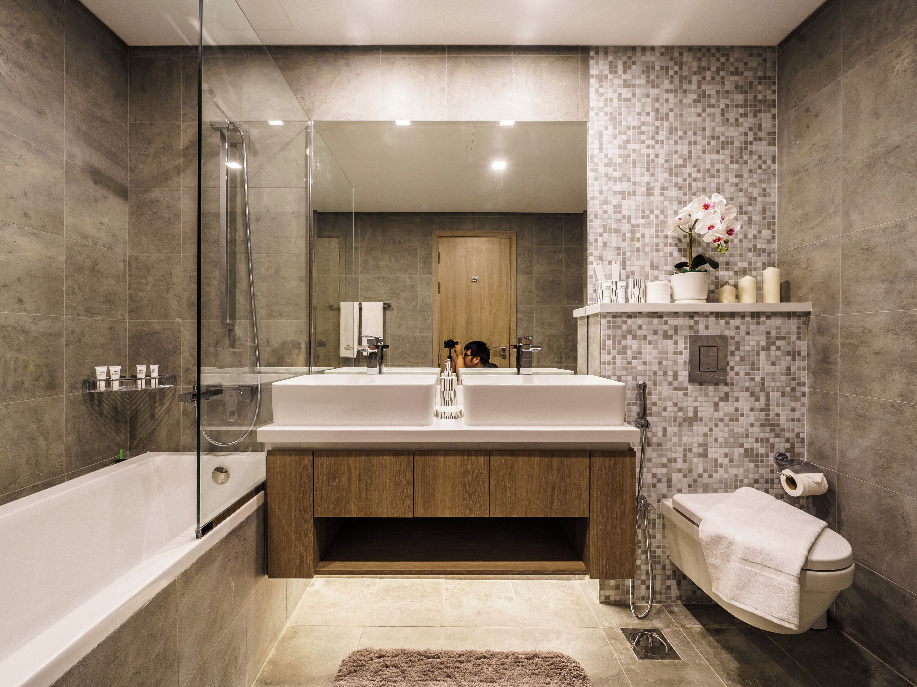 Un bagno moderno con cabina doccia in vetro, un ampio lavandino bianco, mobili contenitori in legno e una parete rivestita in mosaico di piastrelle.