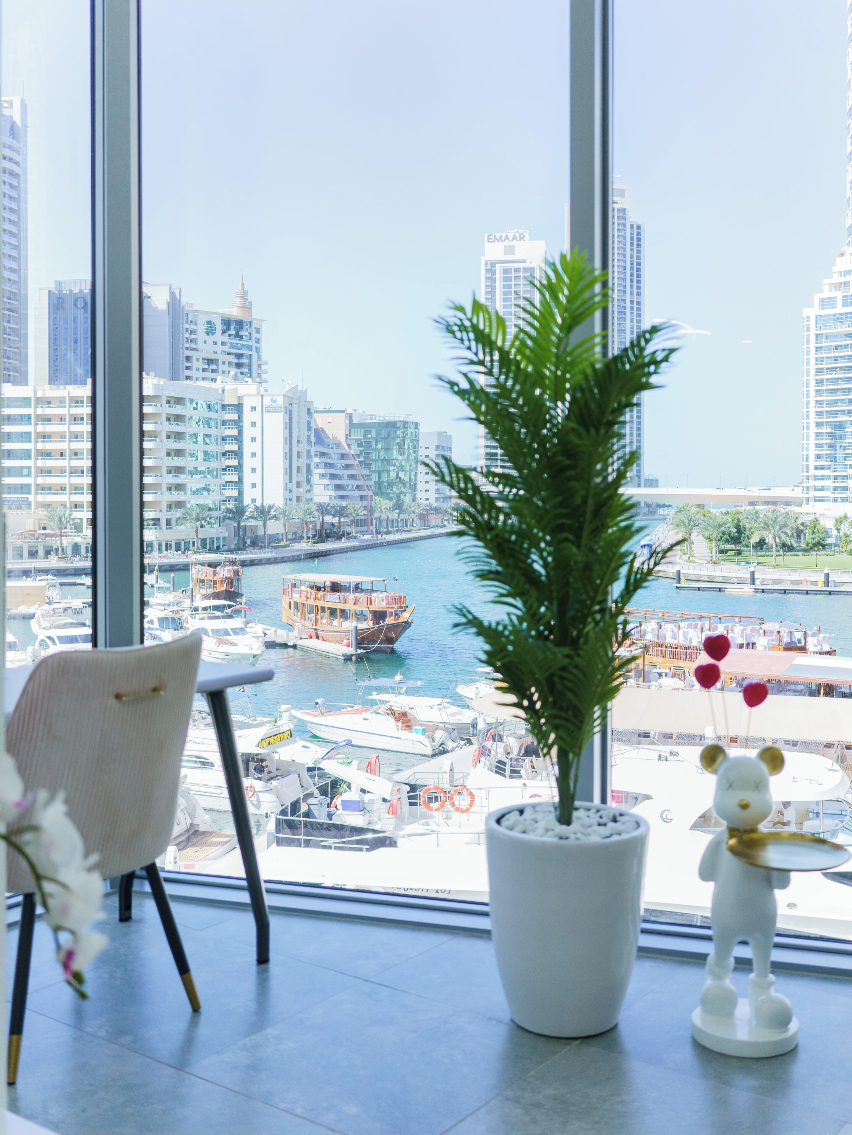 Une vue depuis une fenêtre donnant sur une marina avec des bateaux, le profil urbain d’une ville et un pot de fleurs orné d’une figurine d’ours décorative.