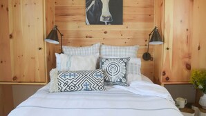 A wooden-paneled room with a bed, two bedside lamps, and a wall-mounted painting. - Tiny House 4 - Farm Sanctuary, Finger Lakes (Watkins Glen)