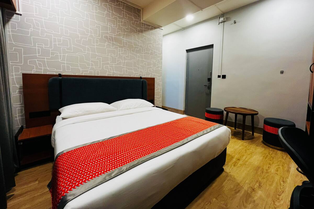 A hotel room with a bed, a wooden headboard, a small table, a stool, and a door.