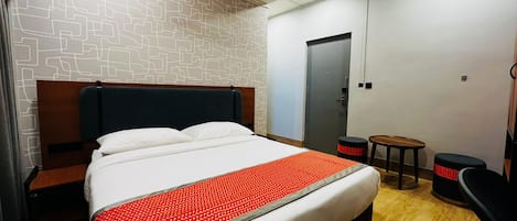 Una habitación de hotel con una cama, cabecera de madera, una mesita, un taburete y una puerta.