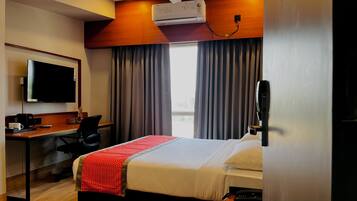 A hotel room with a bed, a desk, a chair, a television, and an air conditioning unit.