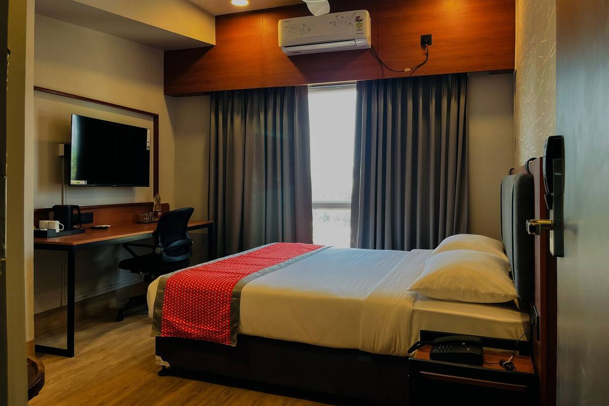 A hotel room with a bed, a desk, a chair, a television, an air conditioning unit, and a window with curtains.