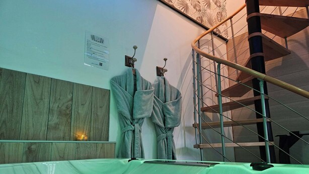 A bathtub with a person in it, a wooden panel wall, a metal spiral staircase, and a robe hanging on the wall. - L'Oxybulles and spa (Friville-Escarbotin)