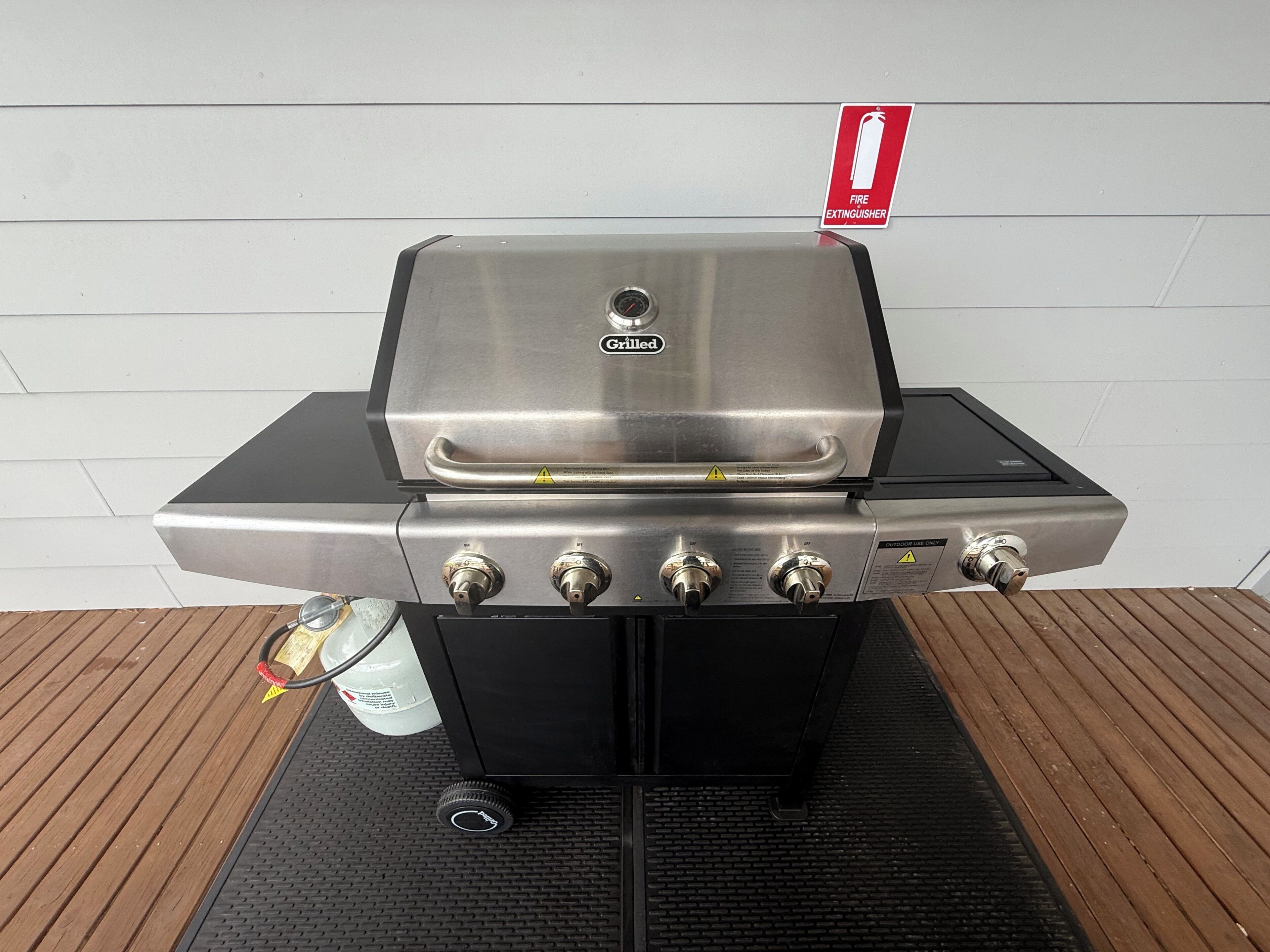 Een roestvrijstalen buitenbarbecue met vier bedieningsknoppen, een bord met daarop een pictogram voor een brandblusser en een aangesloten slang.