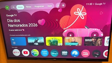 Una pantalla de televisor mostrando la interfaz de Google TV con un tema del 'Dia dos Namorados 2026', que incluye un icono de regalo con forma de corazón y varios iconos de aplicaciones.