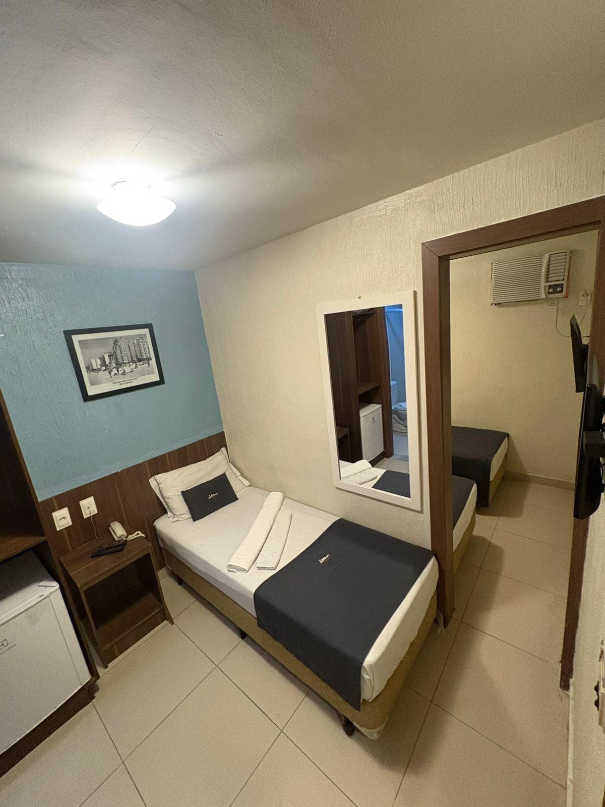 A small hotel room with a single bed, a bedside table with a phone, a mirror, and a picture on the wall.