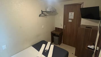 Una habitación pequeña con cama individual, un armario de madera, un televisor montado en la pared y una lámpara en el techo.