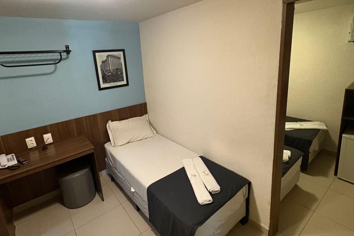 Ein kleines Hotelzimmer mit einem Einzelbett, einem Schreibtisch mit Telefon, einem Papierkorb und einem gerahmten Bild an der Wand.