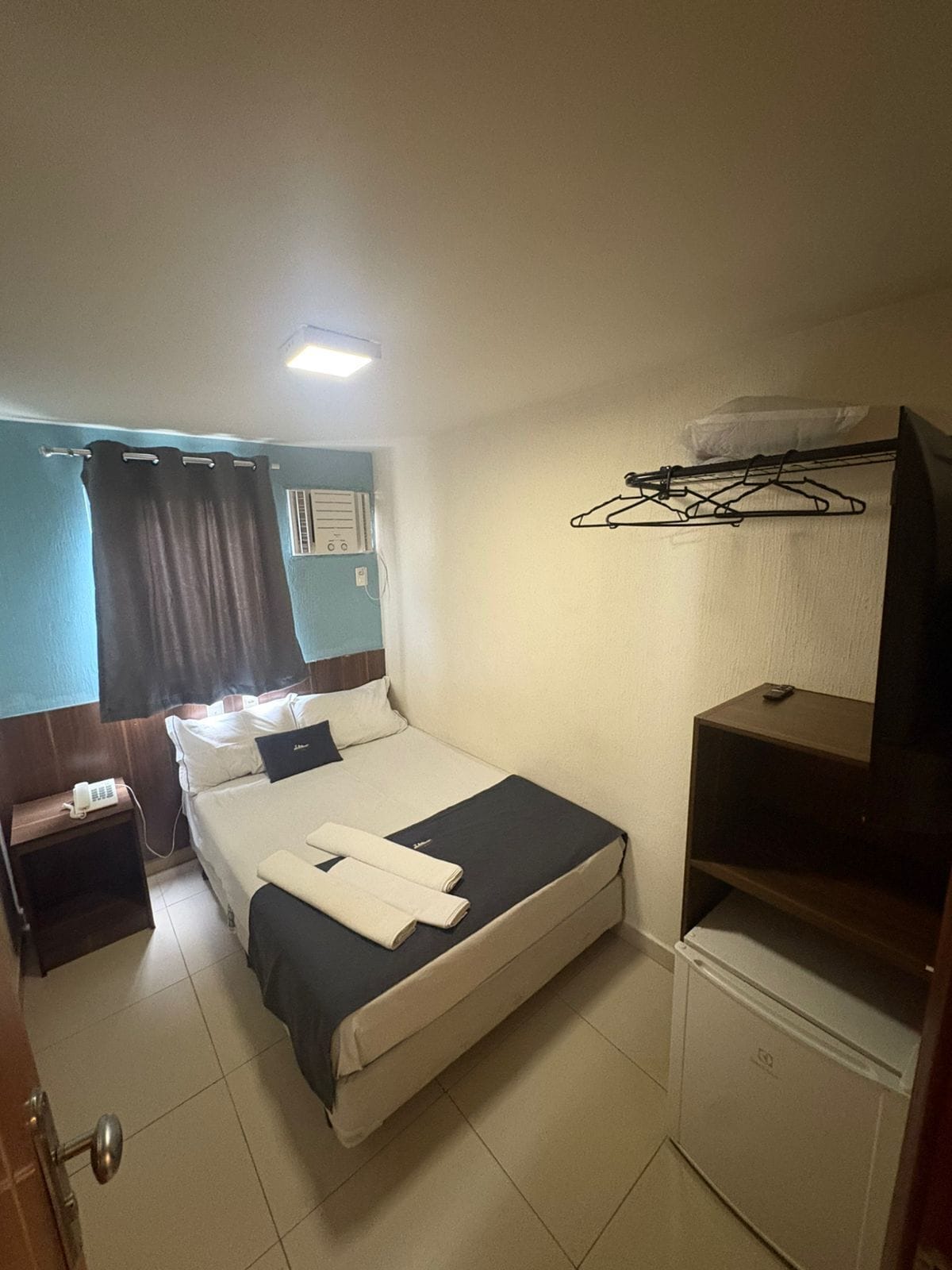 A hotel room with a bed, a bedside table with a phone, a small cabinet, a hanging clothes rack, and a window with curtains.