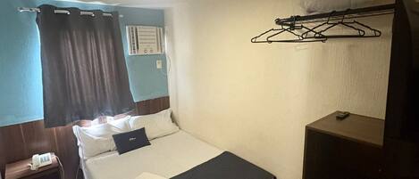 Ein Hotelzimmer mit einem Bett, einem Nachttisch mit Telefon, einem kleinen Schrank, einer Kleiderstange und einem Fenster mit Vorhängen.