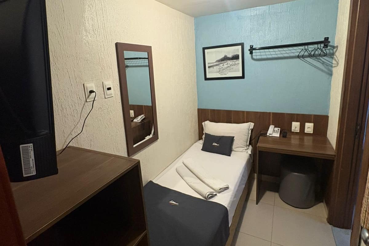 Ein kleines Hotelzimmer mit einem Bett, einem kleinen Schreibtisch, einem Fernseher, einem Spiegel und einem wandmontierten Wäscheständer.