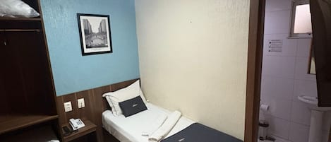 Una cama individual con colchón blanco y manta azul oscuro, una mesita de noche con un teléfono, un estante con un electrodoméstico blanco, y un cuadro en la pared.