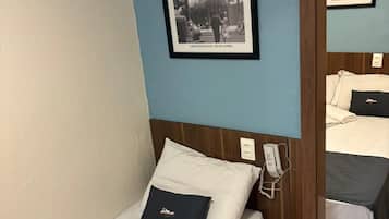 A hotel room with a single bed, a bedside table, a phone, a framed picture on the wall, and a mirror reflecting part of the room.