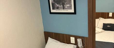 A hotel room with a single bed, a bedside table, a phone, a framed picture on the wall, and a mirror reflecting part of the room.