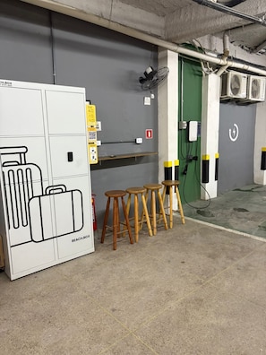 Un cuarto de servicio con un armario blanco grande con la etiqueta 'BEACH.BOX', paneles eléctricos, una puerta verde y una fila de taburetes de madera.