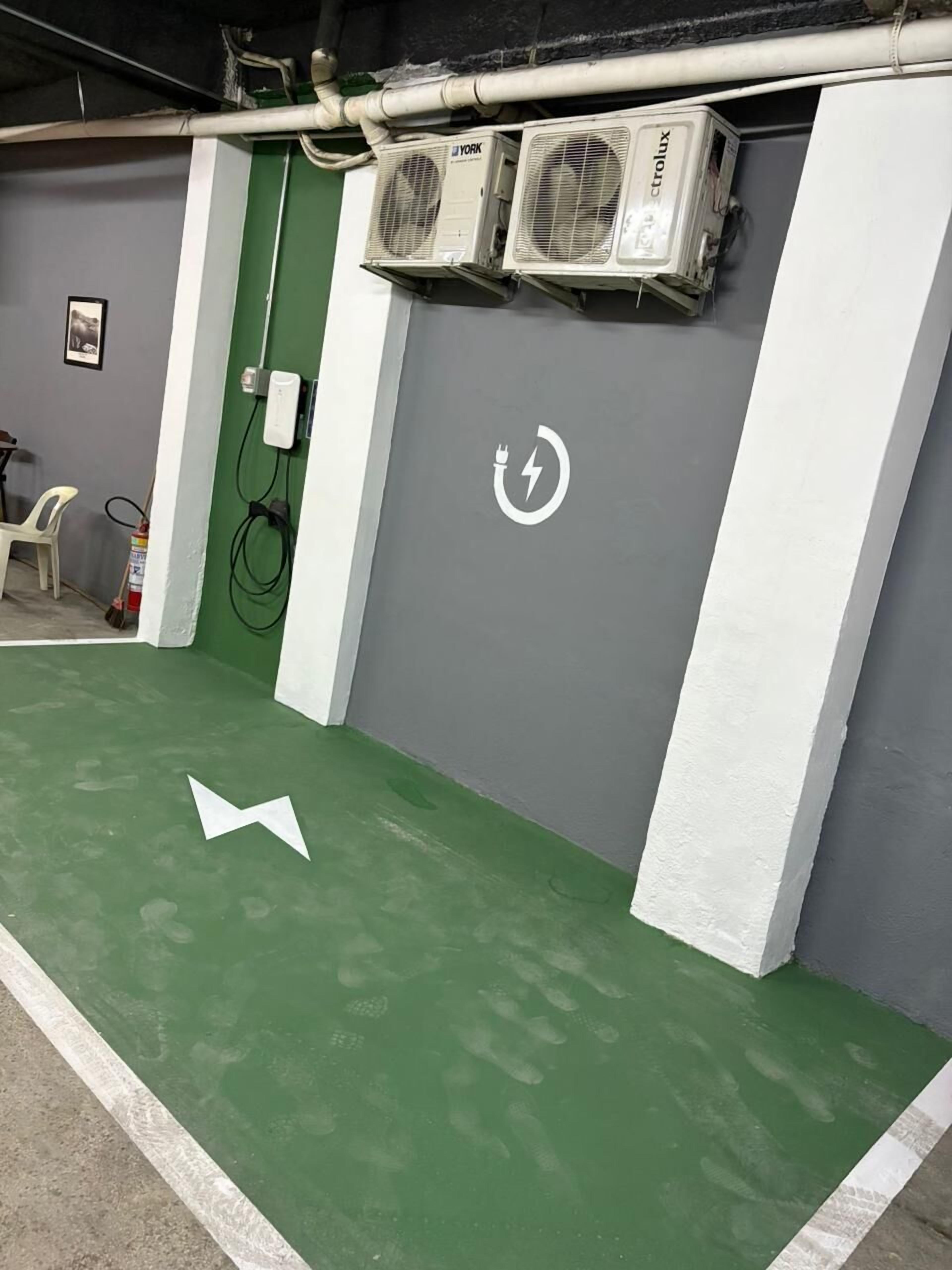 A room with green flooring, white walls, and a grey door. There are two air conditioning units mounted on the wall, a white electrical outlet, and a white arrow on the floor indicating direction.