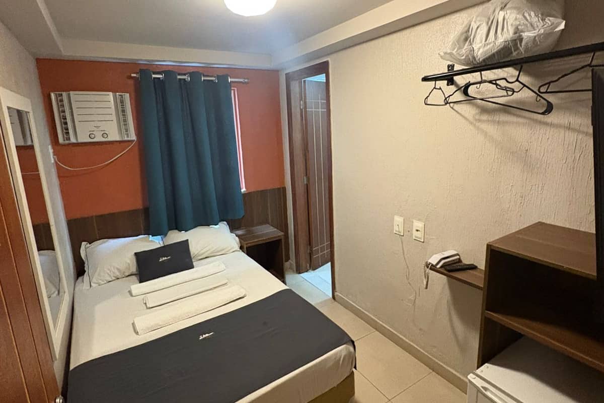 Ein Hotelzimmer mit Bett, kleinem Schreibtisch, Kühlschrank, Heizung, Wandtelefon und Kleiderhaken mit Plastiktüte.