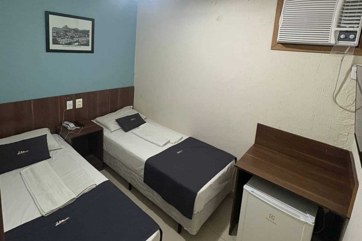 Ein Hotelzimmer mit zwei Betten, einem kleinen Schreibtisch, einem Kühlschrank, einem gerahmten Bild an der Wand und einer Deckenleuchte.