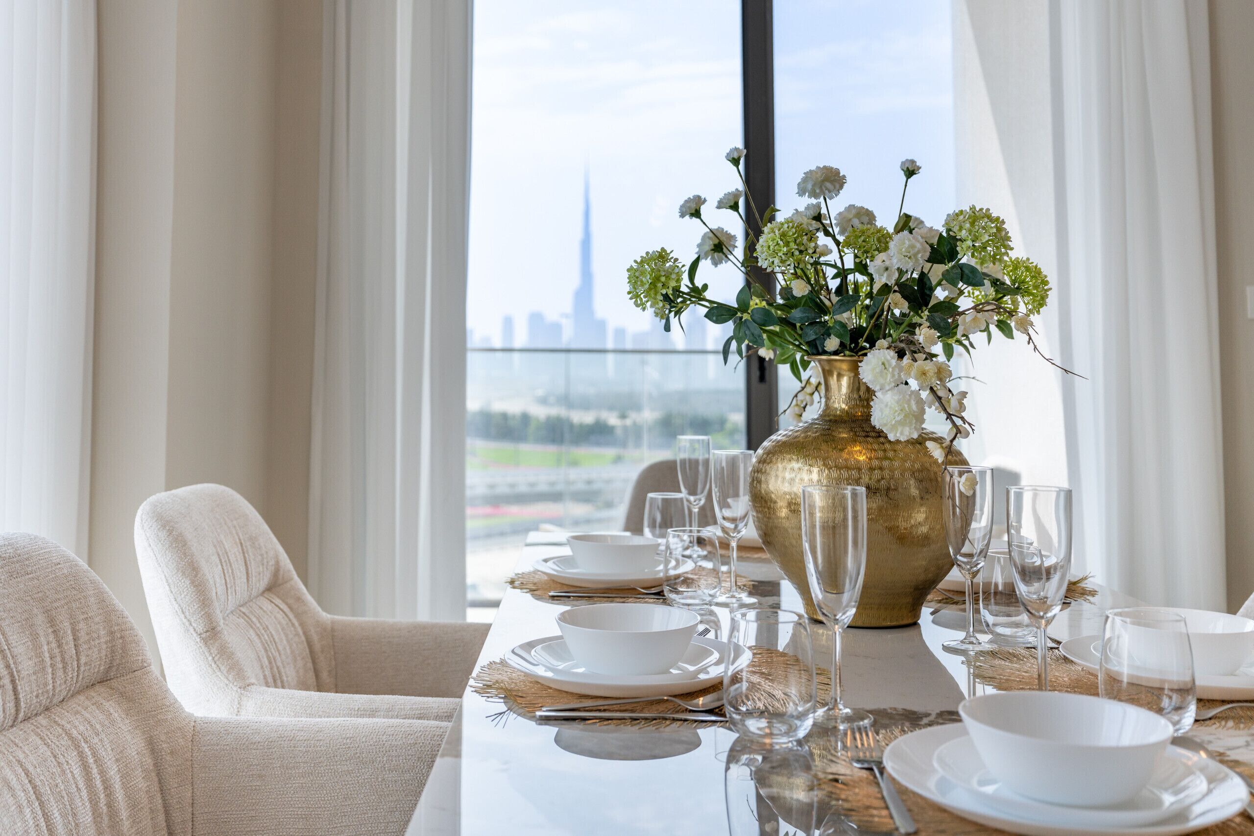 A dining table set with white plates, gold cutlery, and a gold vase with white flowers, with a view of a city skyline through large windows.