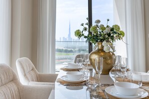 Une table à manger dressée avec de l’argenterie dorée, des assiettes blanches et un vase doré contenant des fleurs blanches, le tout offrant une vue sur les gratte-ciel d’une ville, visible à travers de grandes fenêtres.