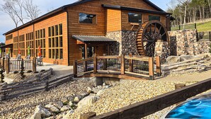 Ein Holzhaus mit großen Fenstern, einem Wasserrad und einem Poolbereich.