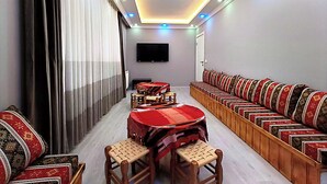 A room with a dining table, chairs, a sofa, a TV, and a ceiling light fixture. - Koçi Hotel (Pazar)