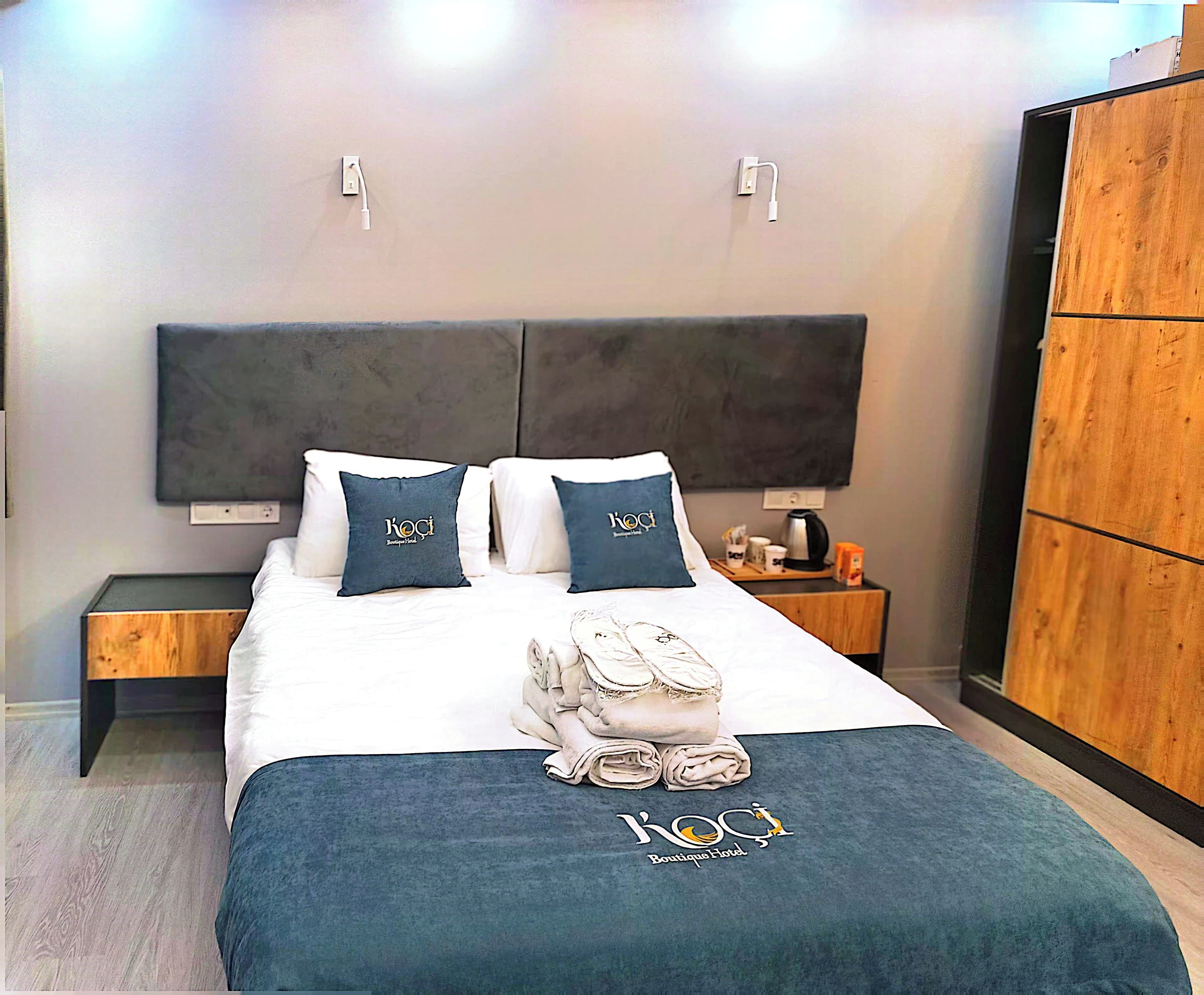 A hotel room with a bed, two pillows with the hotel logo, a bedside table with a kettle, and a wooden wardrobe.