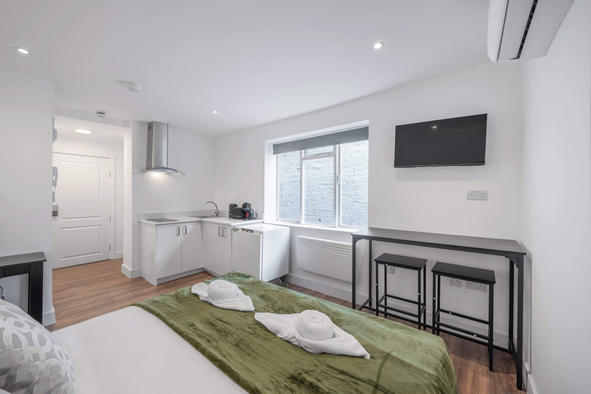 A compact hotel room with a bed, a green blanket, a small kitchenette, a window, a TV, and a desk with two stools.