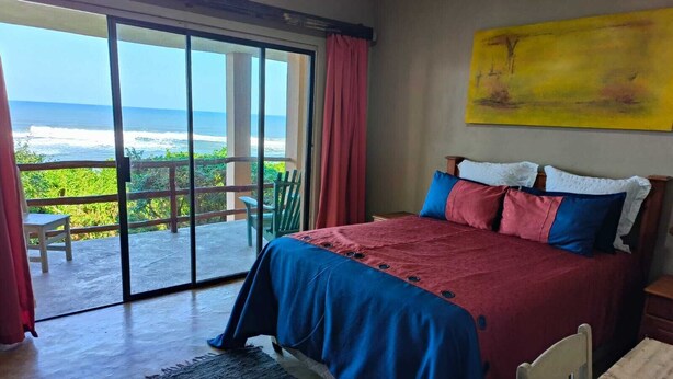 Een slaapkamer met een groot bed, uitzicht op de oceaan, een schilderij aan de muur en een schuifdeur van glas.
