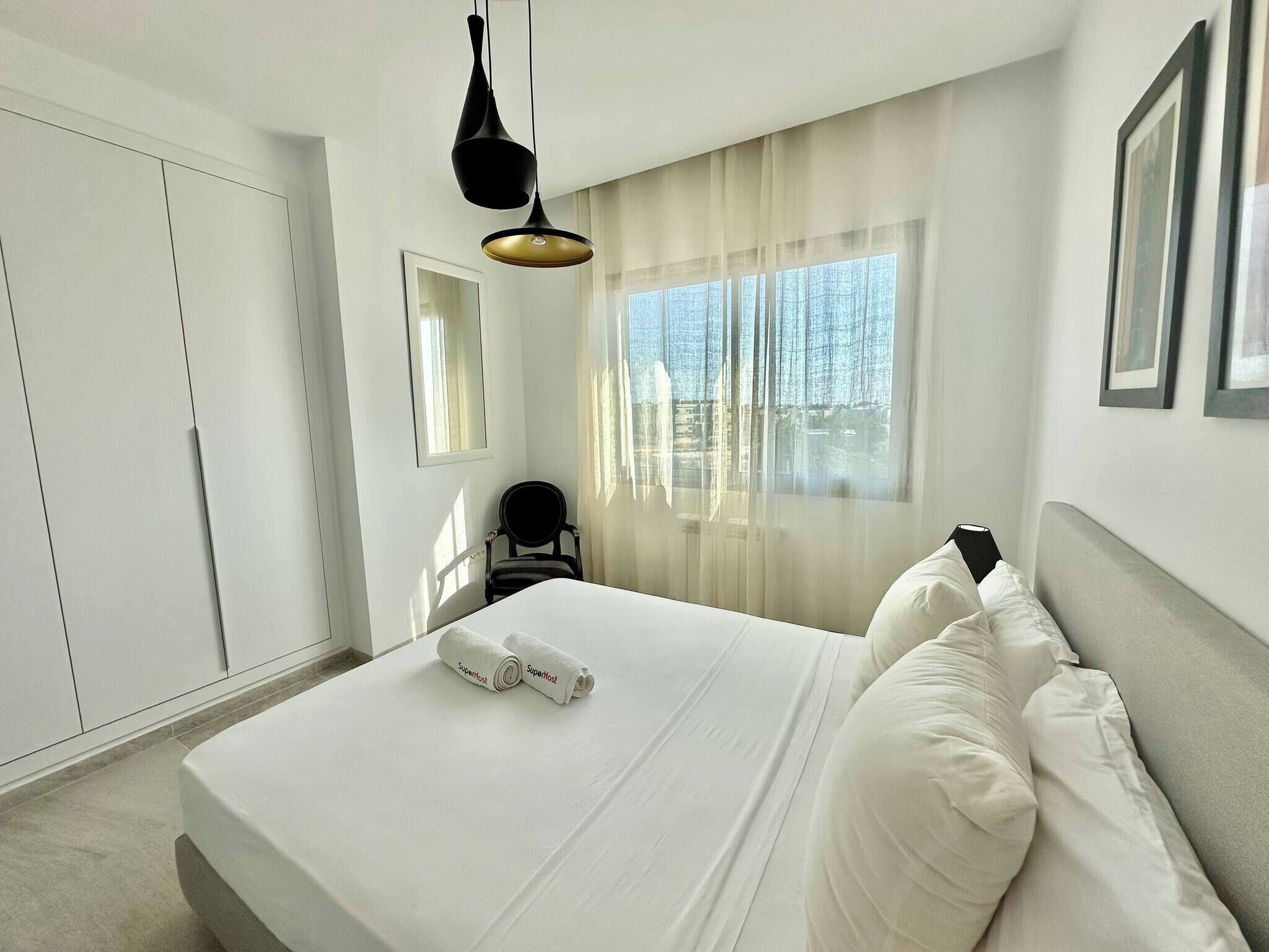 A hotel room with a bed, a chair, a desk, a window with curtains, and a hanging light fixture.