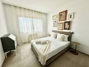 Ein Schlafzimmer mit Bett, Schreibtisch und einem Fenster mit transparenten Vorhängen.