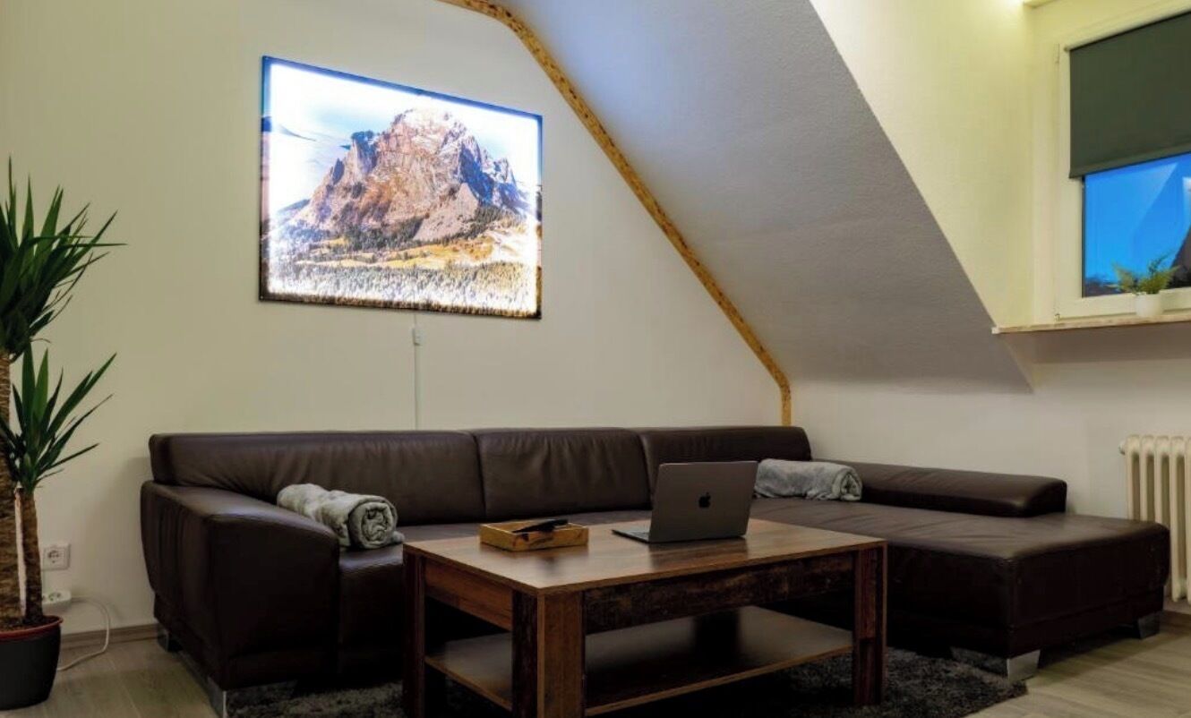 Ein Loft-Wohnzimmer mit schrägem Dach, einem braunen Ledersofa, einem Holztisch mit einem Laptop und einem großen gerahmten Bild an der Wand.
