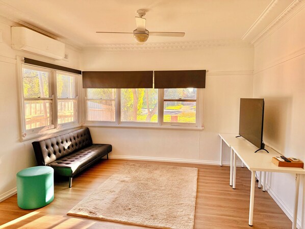 A room with a ceiling fan, a bench, a TV on a stand, a large window with a view of trees, and a green ottoman.