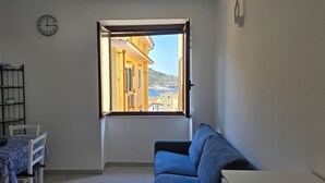Una habitación con una mesa de comedor, sillas, un sofá, un ventanal con vistas a edificios y una masa de agua, y un reloj en la pared.