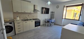 A modern kitchen with a washing machine, stove, sink, and dining area with chairs and a table. - The Sea Cottage (Palinuro)