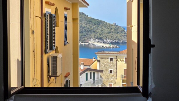 Vista desde una ventana con vista a un pueblo costero, edificios, una masa de agua y una colina al fondo.