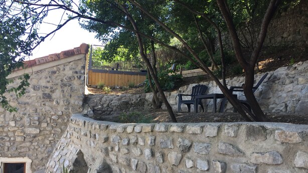 A stone wall with a wooden bench and a chair, surrounded by greenery and trees. - Loft San Francisco (Baix)